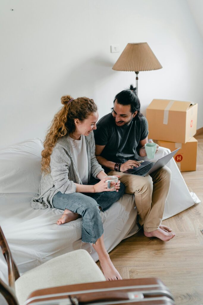 Joyful couple using a laptop and coffee in their new apartment, surrounded by moving boxes.